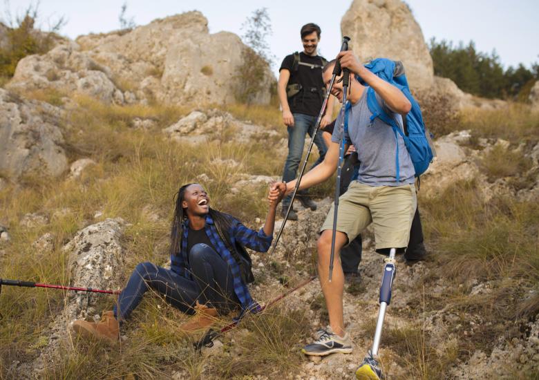 Trois jeunes font une randonnée. Le jeune homme ayant une prothèse de jambe aide une jeune femme à se relever du rocher